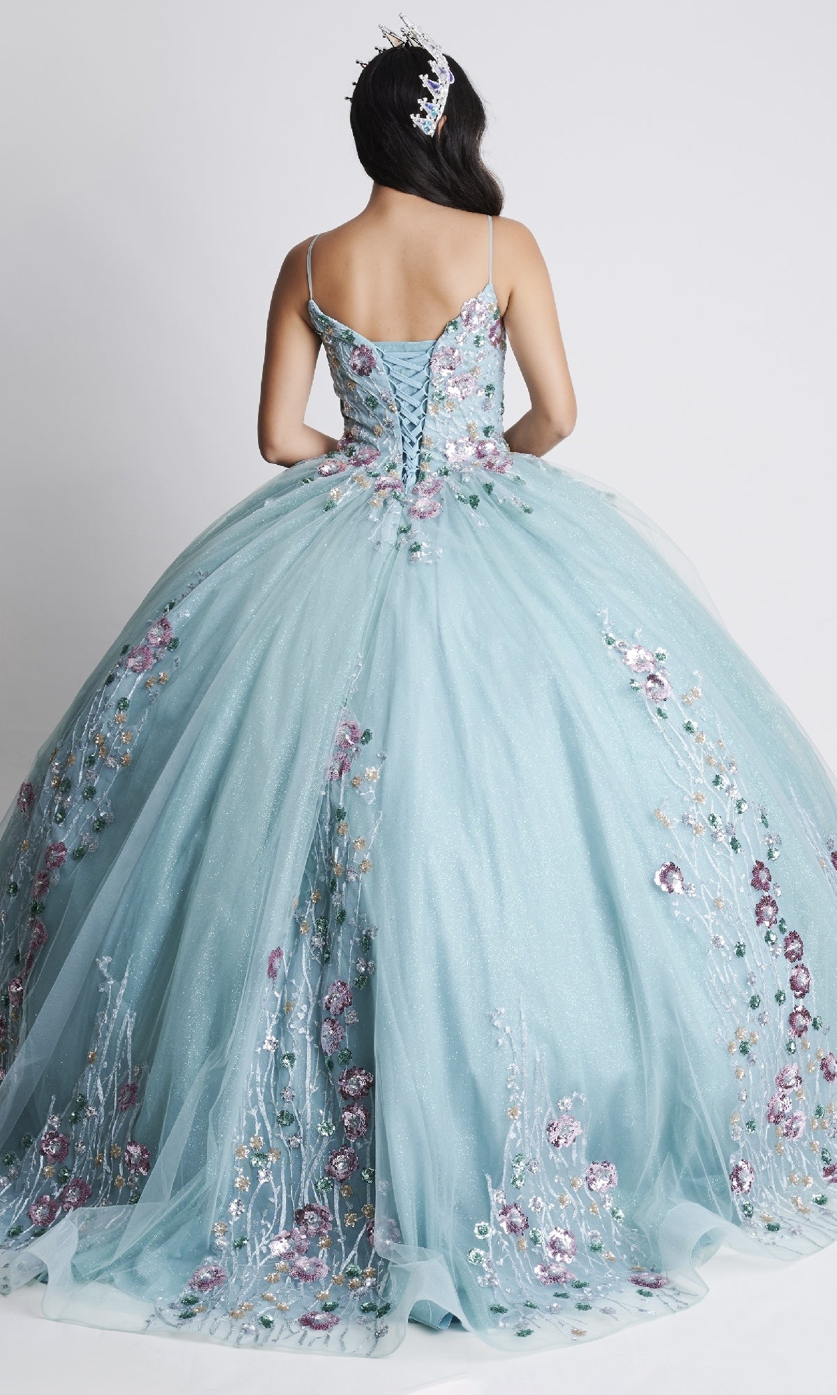 A woman faces away, displaying the intricate floral embroidery and corset back of the Nina Canacci Q105 long Quinceanera dress in light blue, her gown and headpiece adorned with elegant floral accents.